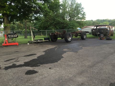 1979 Ford Bronco - Two vehicle frames, one with wheels and tires, sit disassembled in a driveway next to an engine block on a stand.