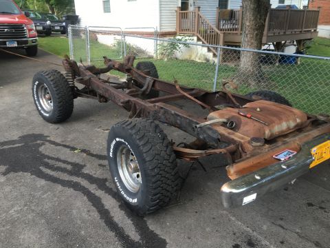 1979 Ford Bronco - The chassis of a vehicle, showing the frame, fuel tank, suspension, and tires, sits on a paved surface.