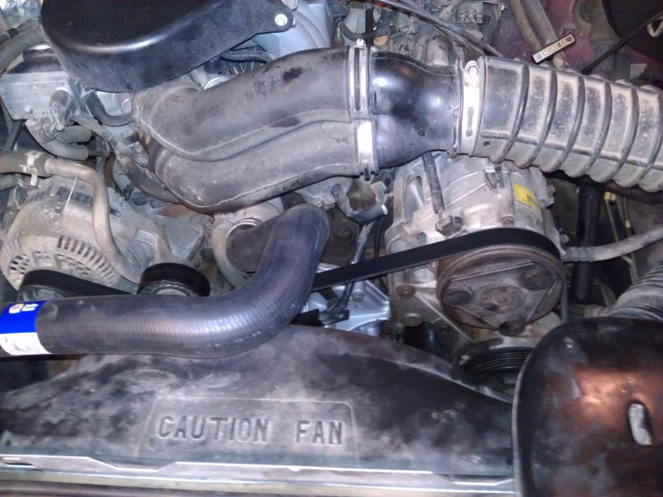 Engine bay with alternator, AC compressor, and fan shroud. Text on shroud reads 'CAUTION FAN'