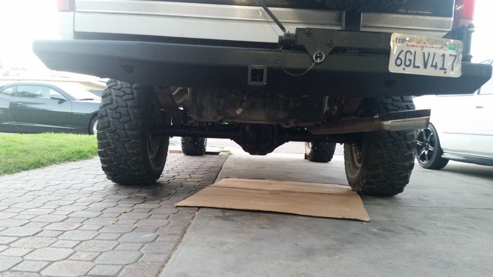 Rear view of a lifted SUV with large tires, aftermarket bumper, and trailer hitch. Another car is visible to the left