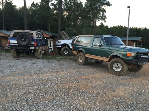 1989 Ford Bronco - Three Ford Broncos are parked in a gravel lot with cabins and trees in the background.