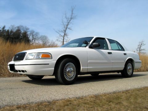2000 Ford Crown Victoria - A white 2000 Ford Crown Victoria sedan is parked on the side of a road with dry grass and trees in the background.