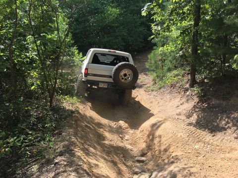 1989 Ford Bronco - White 1989 Ford Bronco navigates a challenging off-road trail with a spare tire mounted on the rear.
