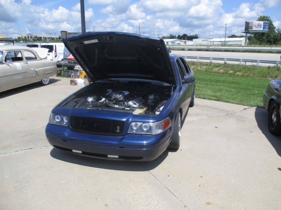 Blue 2004 Ford Crown Victoria with hood open showing the engine bay. Another older car is parked to the left