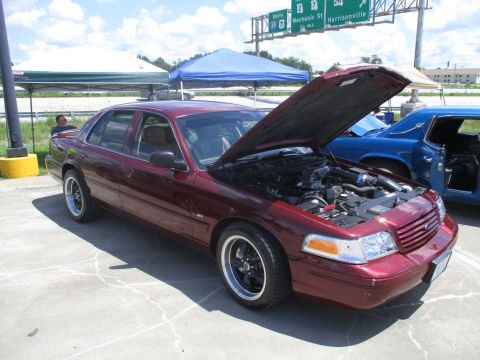 2004 Ford Crown Victoria - A maroon sedan is parked with its hood open, revealing the engine bay, under a bright sky.