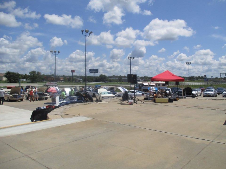Outdoor car show with many cars parked, some with hoods open, under a cloudy sky. Tents and speakers are set up