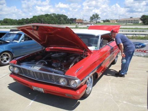 2004 Ford Crown Victoria - A man inspects the engine of a red 1964 Ford Galaxie 500 at a car show, with another car visible in the background.