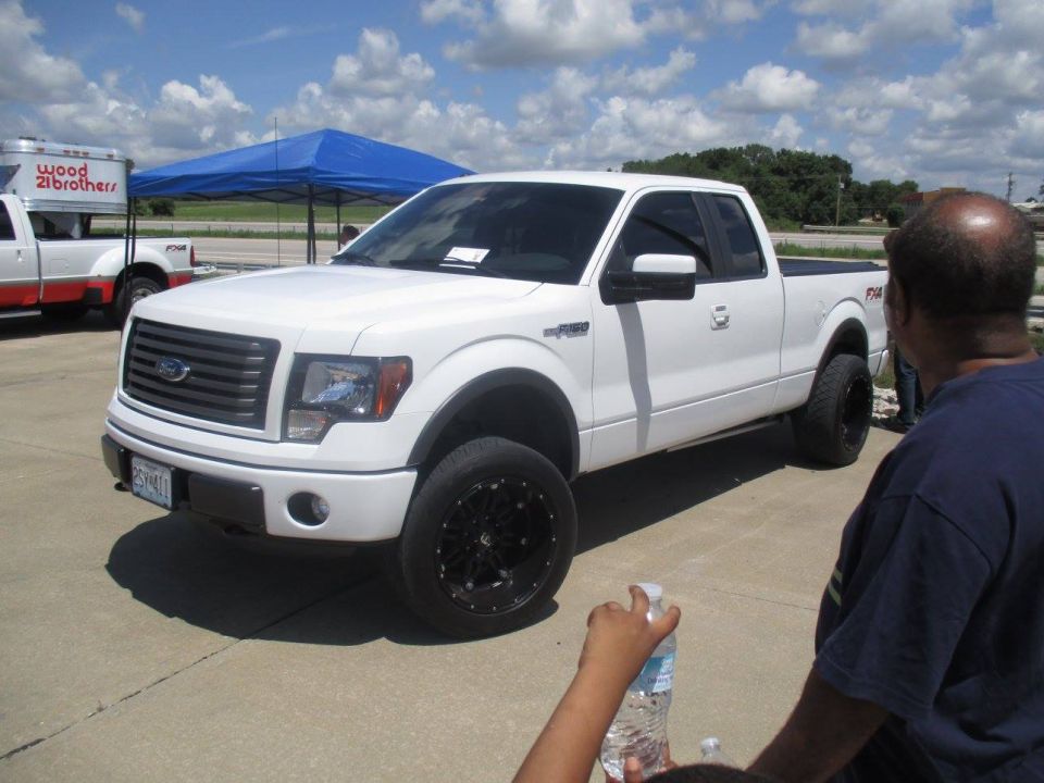 White Ford F-150 pickup truck with black rims and lifted suspension. Another white truck with red trim is parked nearby