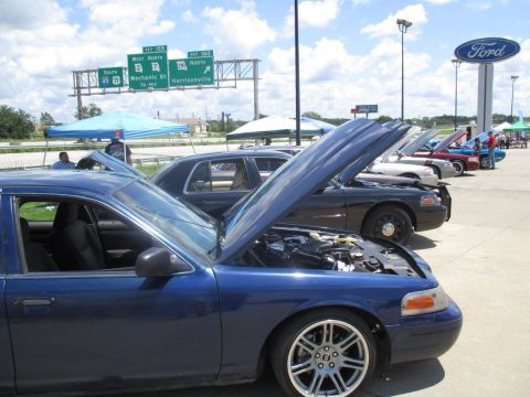 2004 Ford Crown Victoria - Several Ford Crown Victorias are displayed with hoods open at a car event near a highway overpass.