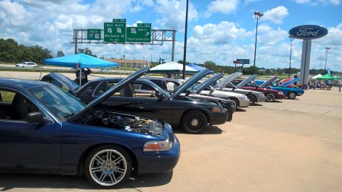 2004 Ford Crown Victoria - A line of Ford Crown Victorias, including a blue one in the foreground, are displayed with their hoods open at an outdoor event.