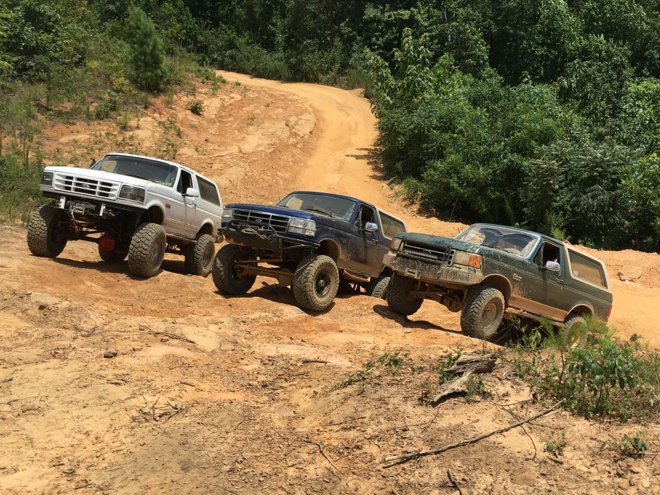Three lifted Ford Broncos driving on a dirt trail with trees on either side