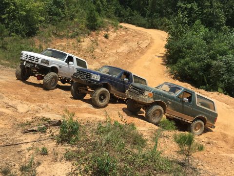 1989 Ford Bronco - Three Ford Bronco SUVs navigate a steep, winding dirt path through a wooded area.