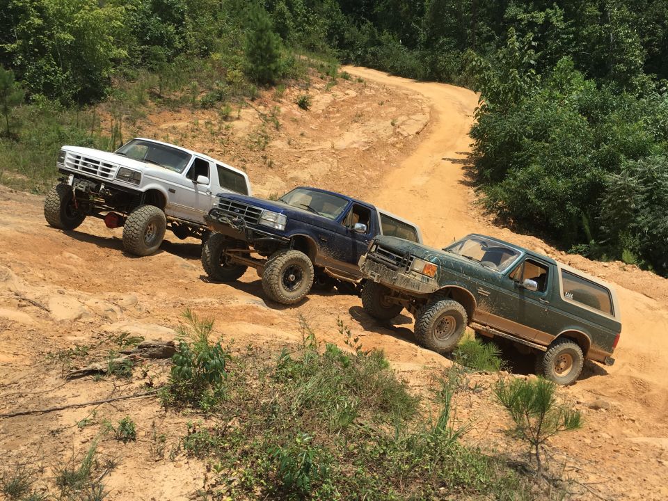 Three Ford Bronco SUVs on a dirt trail, one white, one blue, one green. The green Bronco is in the foreground