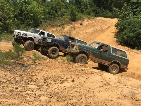 1989 Ford Bronco - Three Ford Broncos navigate a challenging off-road course on a sunny day.