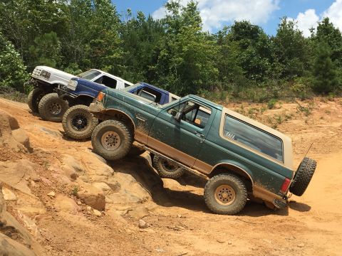 1989 Ford Bronco - Three off-road vehicles navigate a challenging dirt and rock trail under a partly cloudy sky.
