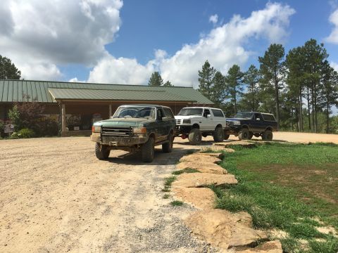 1989 Ford Bronco - Three Ford Broncos, including a green 1989 model, are lined up on a dirt path in front of a building.