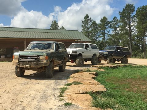 1989 Ford Bronco - A trio of lifted Ford Broncos are parked on a dirt path near a rustic building, with trees in the background.