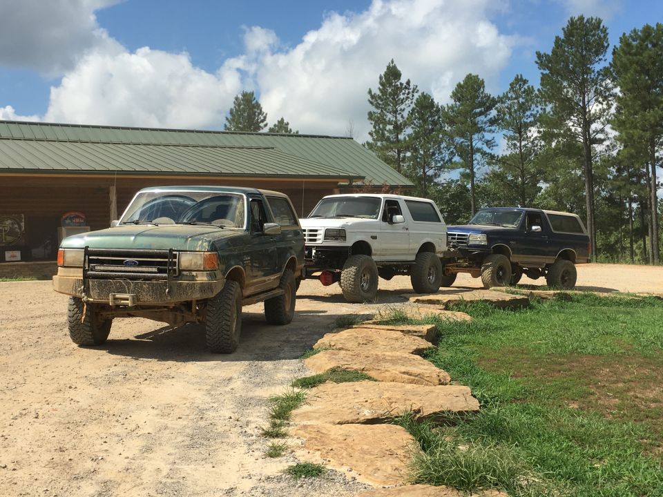 Three Ford Bronco SUVs lined up on a dirt road in front of a building. The first one is green, the second is white, and the third is dark blue