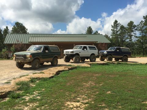 1989 Ford Bronco - Three modified Ford Broncos are lined up in front of a rustic building under a cloudy sky.