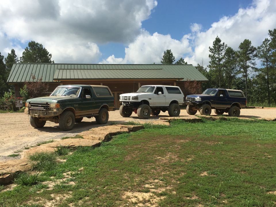 Three Ford Broncos parked in front of a building, one green, one white, one blue, all modified with large tires
