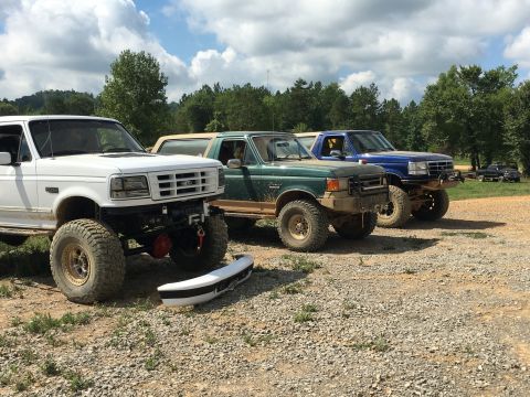 1989 Ford Bronco - Three lifted Ford Broncos, two green and one blue, are parked together on a gravel lot under a cloudy sky.