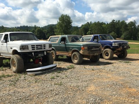 1989 Ford Bronco - Three modified Ford Broncos are parked together outdoors on a gravel lot under a cloudy sky.