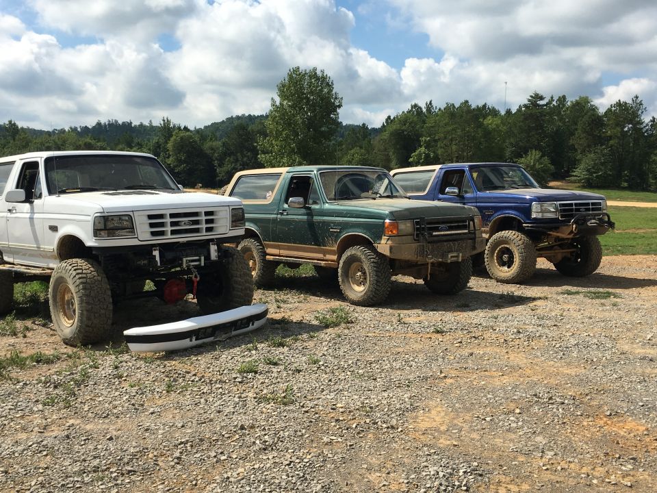 Three lifted Ford Broncos parked on gravel, one white, one green, one blue. The white Bronco has its front bumper removed