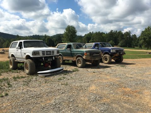 1989 Ford Bronco - Three modified Ford Bronco SUVs are lined up on a dirt and gravel area with trees and hills in the background.