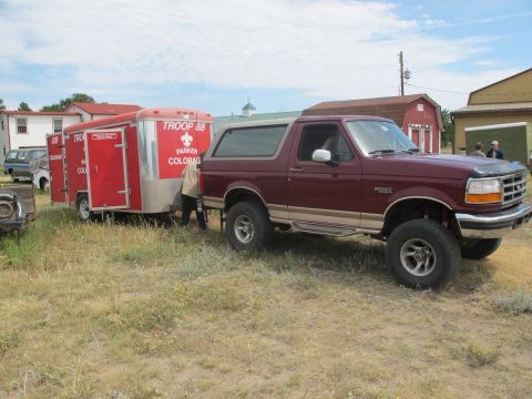 1996 Ford Bronco - A maroon 1996 Ford Bronco is hitched to a red trailer, parked on a grassy area near some buildings.