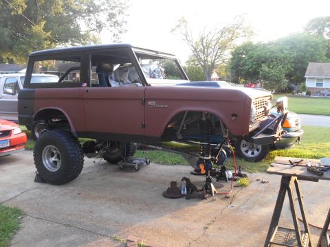 1972 Ford Bronco - A heavily modified 1972 Ford Bronco is shown on a driveway, lifted with its suspension components exposed and being worked on. Various tool…