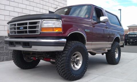 1996 Ford Bronco - A lifted, dark red 1996 Ford Bronco is parked on a concrete surface in front of a brick building. The vehicle features large off-road tires…