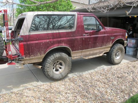 1996 Ford Bronco - The rear passenger side of a muddy, dark red 1996 Ford Bronco is shown parked on a concrete driveway next to a garage. A high-lift jack is…