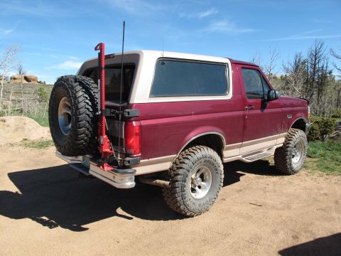 1996 Ford Bronco - The rear passenger side of a maroon 1996 Ford Bronco is shown parked on a dirt road. A spare tire and a Hi-Lift jack are mounted to the rea…