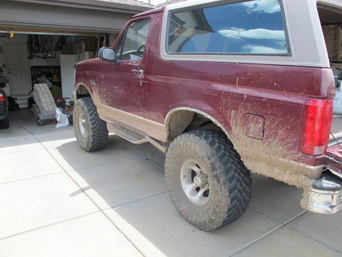 1996 Ford Bronco - The rear driver's side of a mud-splattered, maroon 1996 Ford Bronco is shown parked in a driveway. The vehicle has large off-road tires and…