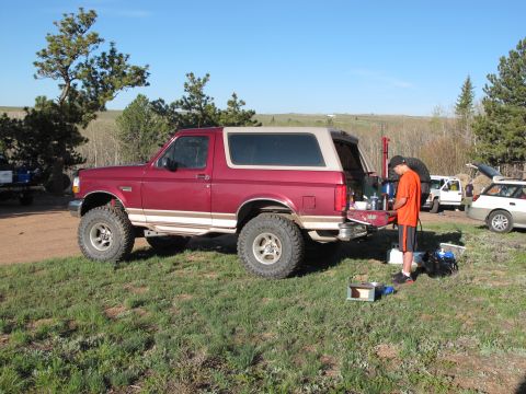 1996 Ford Bronco - A red 1996 Ford Bronco is parked on a grassy area with a person standing next to its open tailgate, preparing food. Another white SUV and a…