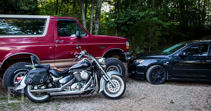Red 1990s Ford Bronco, black sedan, and cruiser motorcycle parked on gravel.