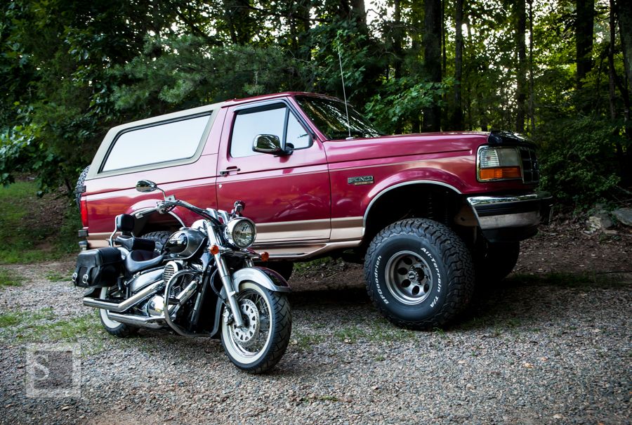 Red 1990s Ford Bronco parked next to a chrome motorcycle in a wooded area.