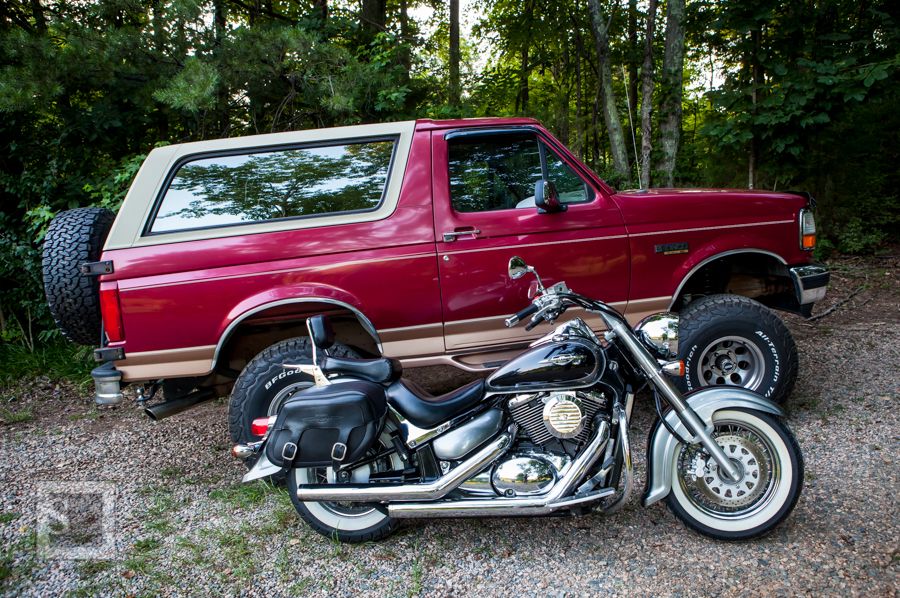 Red 1990s Ford Bronco with tan top parked next to a black cruiser motorcycle with saddlebags.