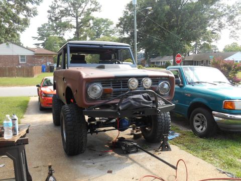 1972 Ford Bronco - Brown early Ford Bronco on lift with front bumper removed in a driveway, teal Ford Ranger parked alongside.