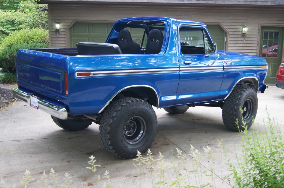 Lifted blue 1979 Ford Bronco, rear 3/4 view, parked in front of a garage.