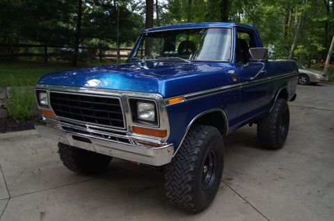1979 Ford Bronco - A blue 1979 Ford Bronco is parked on a concrete driveway in front of a wooded area. The truck has large off-road tires and a chrome bumper…