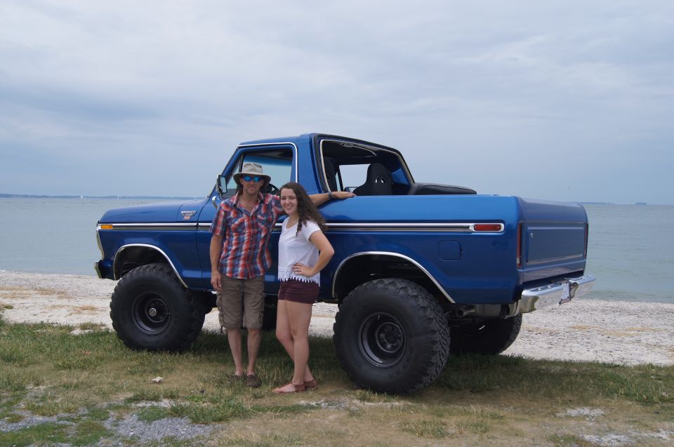 Bright blue lifted Ford Bronco with large tires, a man and a woman posing next to it by the ocean.