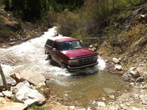 1996 Ford Bronco - A red Ford Bronco is driving through a shallow, rocky stream in a wooded area. The vehicle is partially submerged in the water, creating sp…
