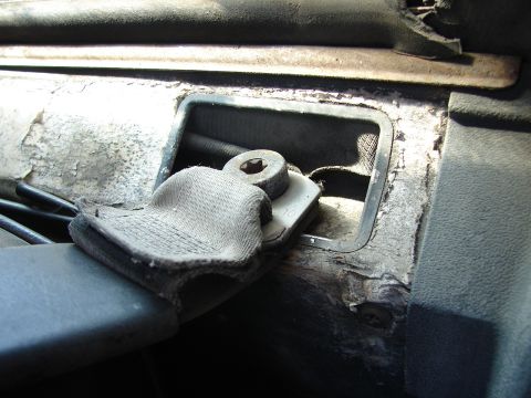 1973 Ford Ranchero - A close-up view of a worn seatbelt buckle mechanism and fabric strap, attached to a dark interior panel of a vehicle. The surrounding area…