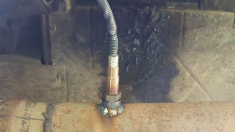 1978 Ford Bronco - A close-up view shows an oxygen sensor threaded into a rusty exhaust pipe, with a black cable extending to the left.