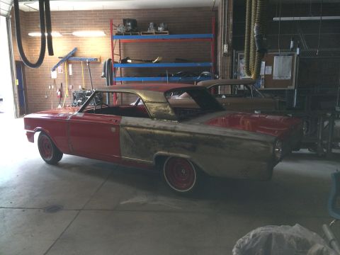 1964 Ford Fairlane - A red 1964 Ford Fairlane coupe is in a workshop, with one side showing bare metal bodywork, indicating it is undergoing restoration.