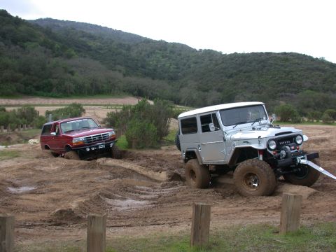 1996 Ford Bronco - Two off-road vehicles navigate a muddy terrain with hills in the background.