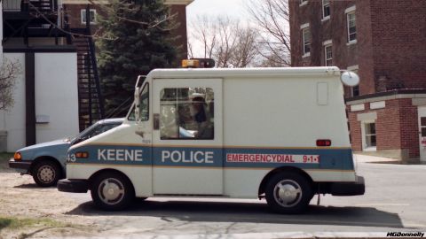 1991 Ford Crown Victoria LTD - A Keene Police van is parked next to a blue car, with buildings in the background.
