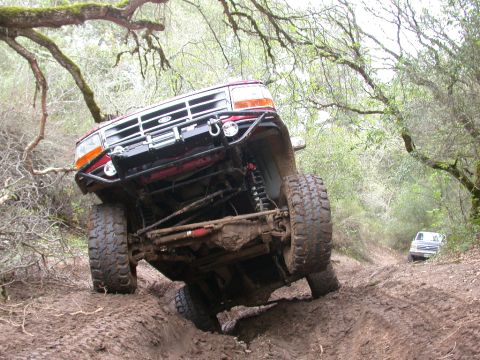 1996 Ford Bronco - A red 1996 Ford Bronco navigates a challenging muddy trail, with another vehicle visible in the distance.