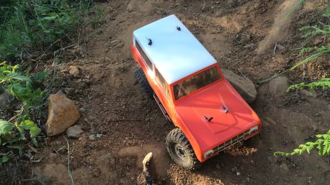 1989 Ford Bronco - RC rock crawler toy with orange body and white roof navigates a dirt trail.